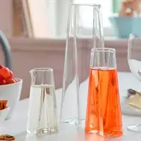Tre vinklet glas kander; en tom, en med klar væske, en med orange væske, på bord med frugt.